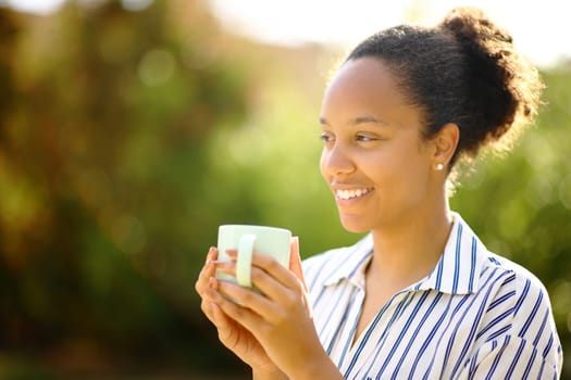 Happy black woman holding coffee mug contemplating views in a park