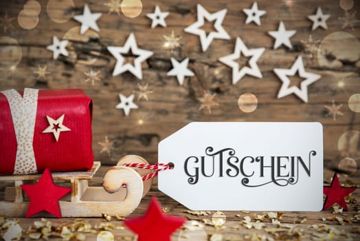 Wooden Sled With Red Gift And Rustic Christmas Bokeh Background With Label With German Text Gutschein, Which Means Voucher In English