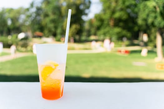 A glass with an Aperal Spritz cocktail, a slice of orange and a straw in the park in summer.