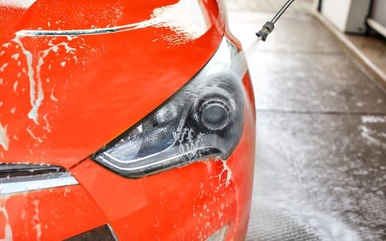 Front light of red car washed in self serve carwash, jet water spraying on glass.