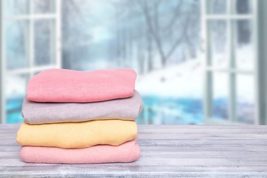 Stack of winter clothes. A pile of colorful cozy warm sweaters on a wooden table against abstract blurred natural winter background. Autumn and spring clothes. Copy space.
