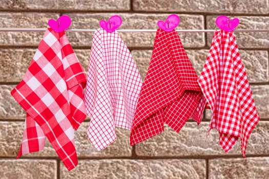 Hanging towels. Closeup of various red checkered kitchen towels with violet pink clothespins hang on a clothes rail against abstract blurred brick wall.