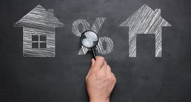 Chalk-drawn houses and a percentage sign on a blackboard, a female hand holding a magnifying glass. Concept of increasing or decreasing mortgage, rent.