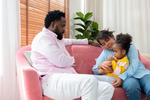 Angry african father scolding his stubborn daughter at home while curly little daughter feels upset. Mother comforting and embracing daughter while sitting together on couch. Family problems and conflicts concept