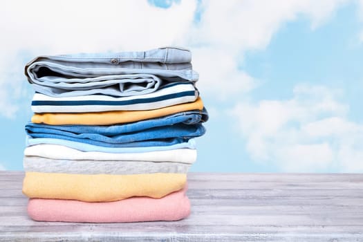Stack of jeans. Closeup of a pile of colorful denim pants and colorful sweater on a bright table against abstract blurred natural light blue sky background. Space for your product display montage.