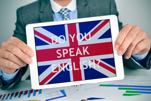 closeup of a young caucasian businessman at his office desk full of charts showing a tablet computer with the question do you speak English? and the flag of United Kingdom in its screen