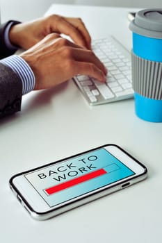 closeup of a smartphone with the text back to work in its screen, on the office desk of a businessman who is typing in the keyboard of his computer