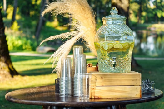 Lemonade in a glass decorative beverage dispenser, one-time glasses on the table during a garden party.