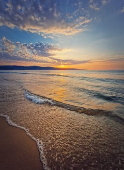 Vibrant sea sunrise with foamy waves washing the sandy beach. Dawn scene with the sun rising up the hills on the horizon
