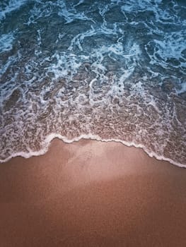 Soft foaming wave of the blue sea hits the sandy shore. Beach and water texture, moody vertical background. Summer vacation seaside, holiday recreation concept