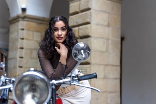 Attractive young woman with long black  hair is posing by historic motorcycle. Horizontally.