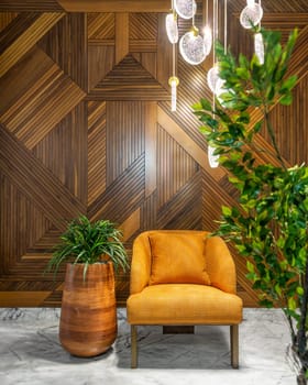 Modern orange armchair, tall wooden planter, and contemporary glass chandelier, in a hall with decorated wood cladding wall, and white marble floor, framed by green bushes