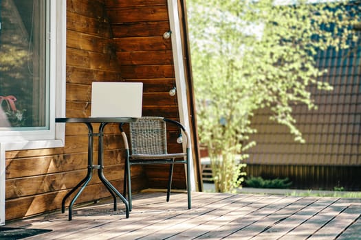 Laptop on the coffee table near whicker chair on terrace of log cabin. Concept of remote work and freelance.