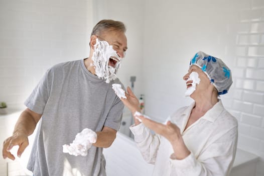 Happy loving senior married couple fooling around in bathroom. Cheerful old husband and wife laughing and playing with shaving foam. Happiness and positivity