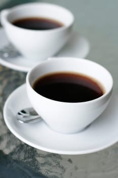 Two black filter coffees in white cups on white saucers on a glass table.