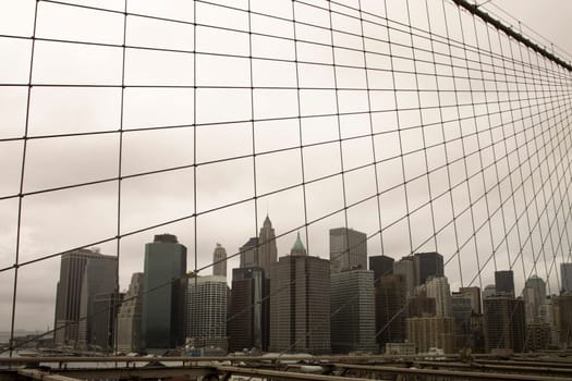 A view of Manhattan downtown and the skyscrapers near Wall Street from the Brooklyn Bridge. The gloomy sky with leaden gray clouds is segmented by a web of cables from the suspension bridge.