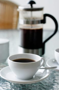 A black filter coffee in a white cup with a coffee plunger in the background on a glass table.