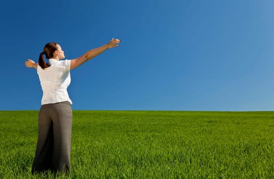A beautiful young woman holding her arms out in praise or celebration standing in a green field and facing towards the light in a blue sky