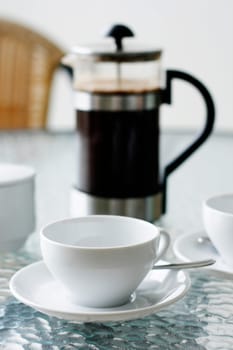 A black filter coffee in a white cup with a coffee plunger in the background on a glass table.