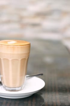 A latte in a glass on a white saucer with basic coffee art design on a café table.