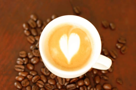 Top down view of a delicious piccolo latte in a small white cup with a heart shaped coffee art design sitting on a wooden table surrounded by roasted coffee beans.