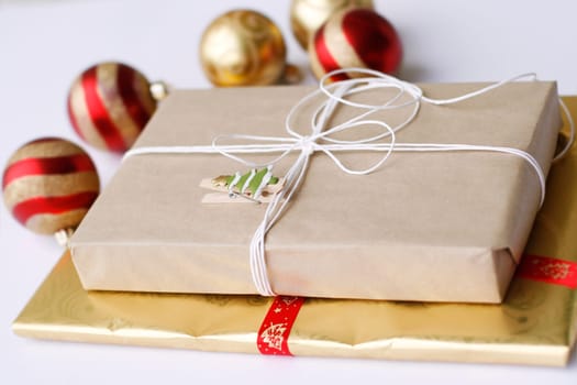 A close-up of a two Christmas gifts that are simply wrapped, one with light colored paper completed with a white bow string and clipped on decoration, and the other with gold colored paper and then, as well as Christmas baubles in the background.