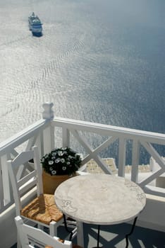 View across a balcony with table and chairs, looking down over the caldera in Santorini, where a cruise ship has arrived