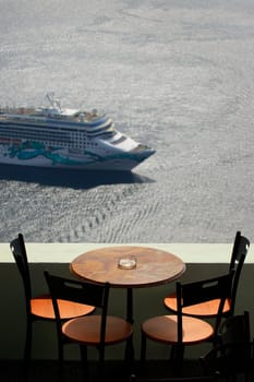 View across a balcony with table and chairs, looking down over the caldera in Santorini, where a cruise ship has arrived