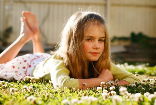A portrait of a pretty and happy long haired young girl posing for the camera while lying on the grass amongst little flowers.