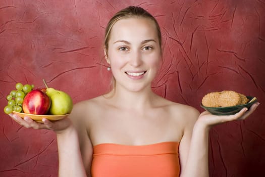 Smiling young woman on a diet. Girl choosing food: fruits or cookies. Red background.