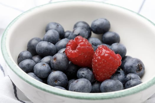 A close-up of fresh blueberries and raspberries in a bowl sitting on a white kitchen cloth.