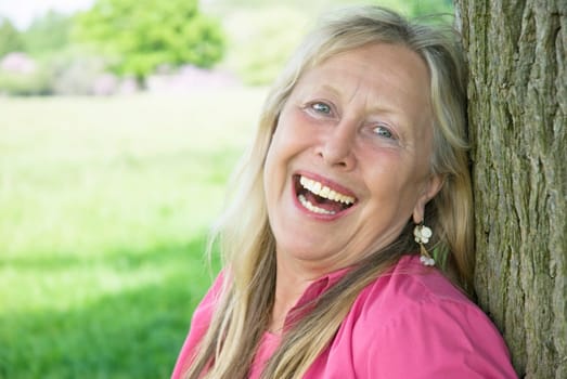 A beautiful older woman in her sixties shows her wonderful smile as she leans against a tree during a day out in the countryside.