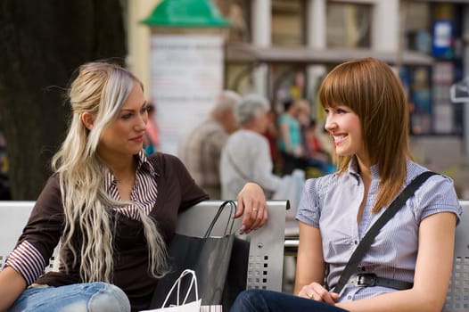 two nice girls sitting on a bench outdoor and chatting each other and smiling