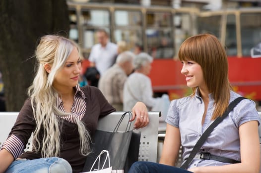 two nice girls sitting on a bench and chatting each other during a shopping day