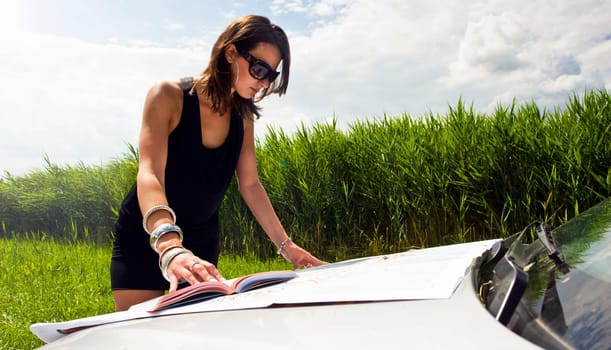 Young woman, studying a map and a guide book on the hood of her car on a bright summer day