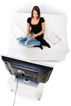 Young woman, sitting comfortably on a white couch with a magazine and the remote next to her, drinking a cup of tea and watching television