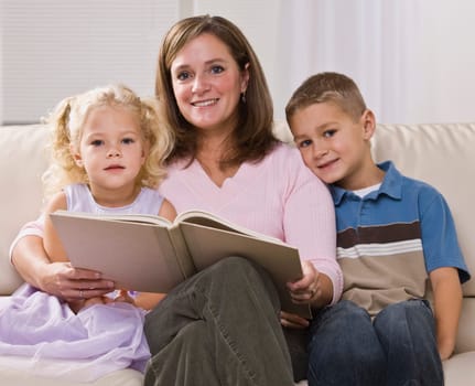 A mother is reading to her two young children.  They are all smiling at the camera.  Square framed shot.