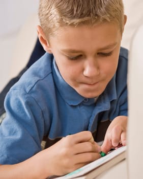 A young boy is laying on his stomache and coloring in a coloring book.  He is looking away from the camera.  Vertically framed shot.