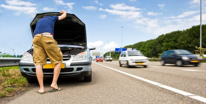 Man staring down at the engine of his stalled car on the hard shoulder of a motorway