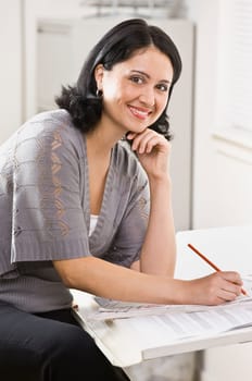 A young, attractive woman is looking over some paperwork on a desk, and smiling at the camera.  Vertically framed shot.