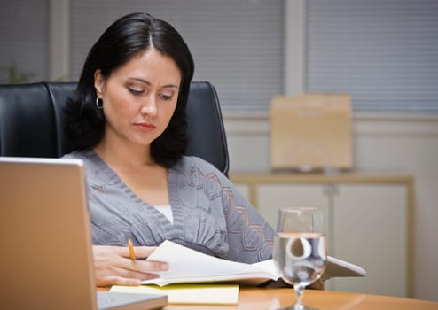 A young businesswoman is seated at a desk in an office.  She is looking away from the camera.  Horizontally framed shot.