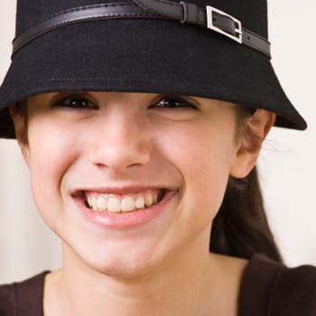 A young girl is wearing a hat and smiling at the camera.  Square composition.