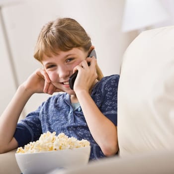 A young girl is seated on a living room sofa and is talking on a cellphone.  She is smiling at the camera.  Square framed shot.