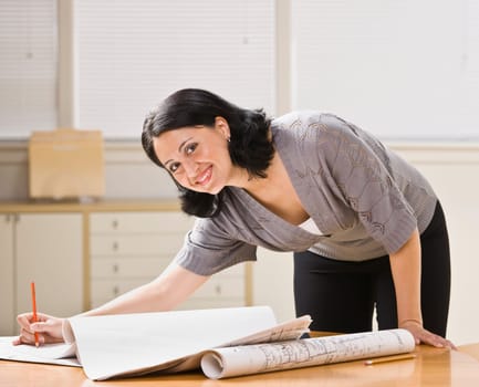 Attractive woman leaning over desk with drawings. Pen in hand, looking at camera. Horizontal.