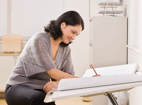 A young woman is sitting at a desk in an office and is working on blueprints.  She is looking away from the camera.  Horizontally framed shot.