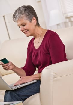 An elderly woman is seated in a couch with a laptop.  She is looking at a card in her hand and smiling.  Vertically framed shot.