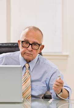 An elderly man is working on a computer in an office.  He is looking at the camera.  Vertically framed shot.