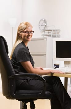 A businesswoman is seated at a computer desk and smiling at the camera.  Vertically framed shot.