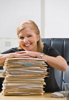A businesswoman is seated at a desk in an office with a stack of paperwork.  She is smiling at the camera.  Vertically
