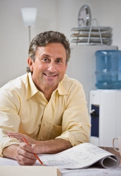 A man is sitting at a desk and working on blueprints.  He is smiling at the camera.  Vertically framed shot.